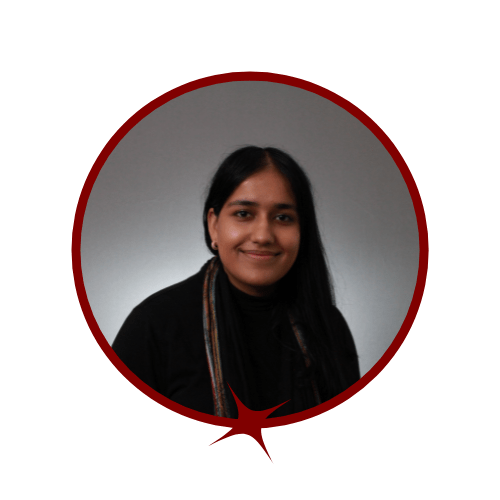 Picture of dark-haired woman wearing a black long-sleeve dress and sitting in front of a grey background. 