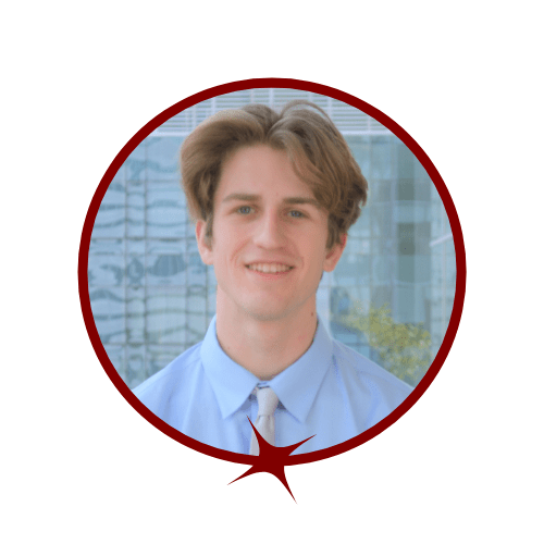 Picture of blond-haired man wearing a blue dress shirt and silver tie. He is standing in front of a tall window. 