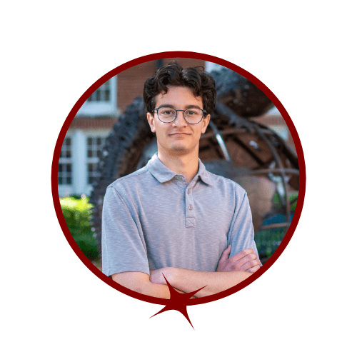 Picture of dark-haired man wearing a light blue shirt standing in front of a tree and gator statue. 