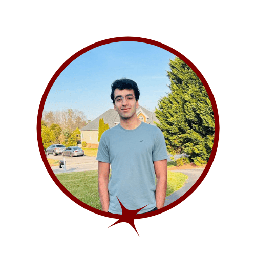 Picture of smiling, dark-haired man wearing a blue shirt standing in front of nature. 