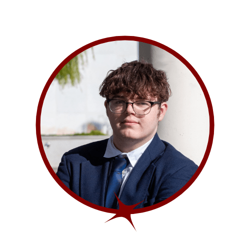 Picture of auburn-haired man wearing glasses and a blue tuxedo blazer in front of a tree background. 