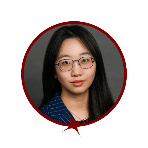 Picture of dark-haired woman wearing glasses and a navy blue blazer standing in front of a grey background. 