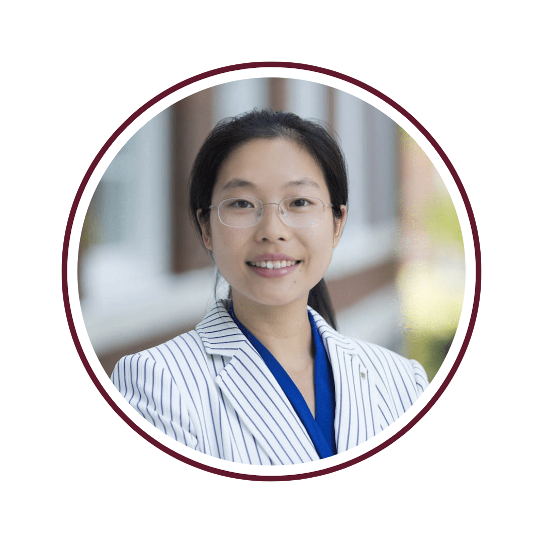Young dark-haired woman standing in front of blurred residential background. She is wearing a blue and white striped blazer with a dark blue undershirt. The picture is bordered by a maroon circle. 