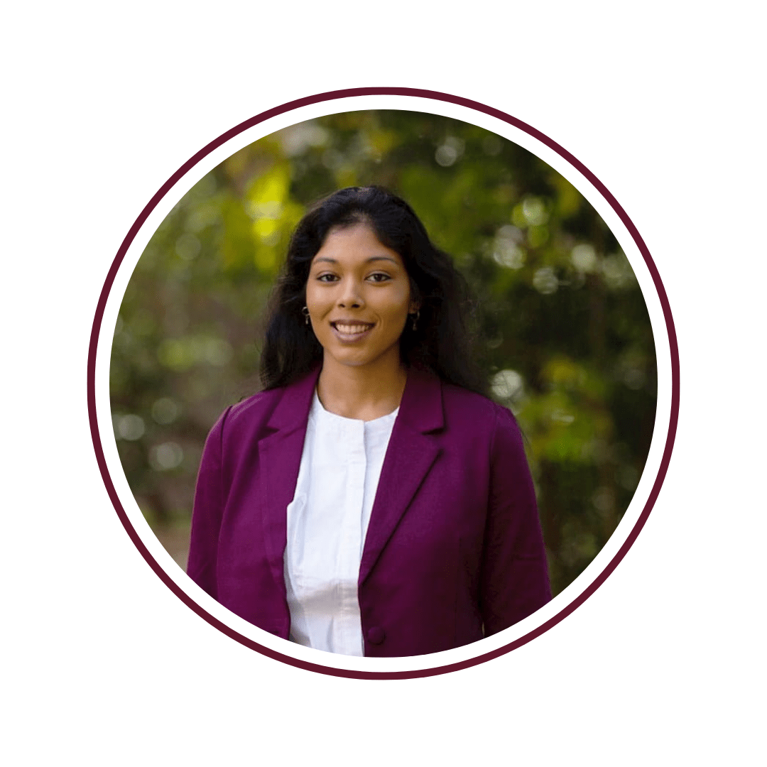 Woman in purple blazer and white shirt stands in front of blurred green background. The picture is framed by a maroon circle border. 