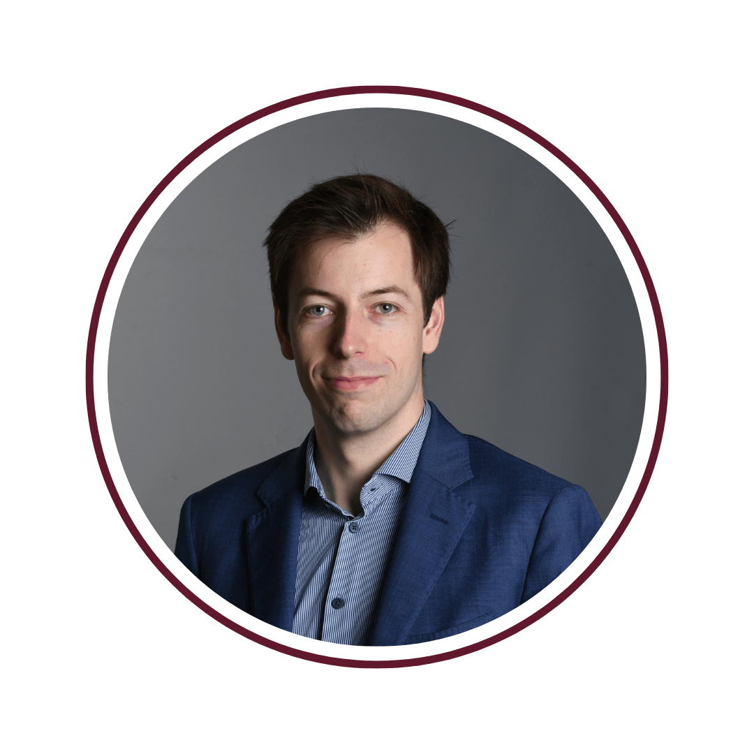 Young smiling man standing in front of gray background. He is wearing a navy blue blazer and light blue undershirt. The frame is a maroon circle. 