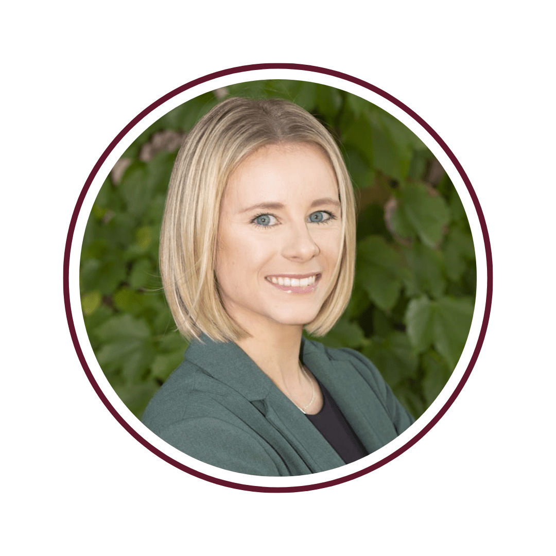 Young blonde woman with blue eyes standing in front of a green vine background. She is wearing a grey blazer and black undershirt. The picture is bordered by a maroon circle. 