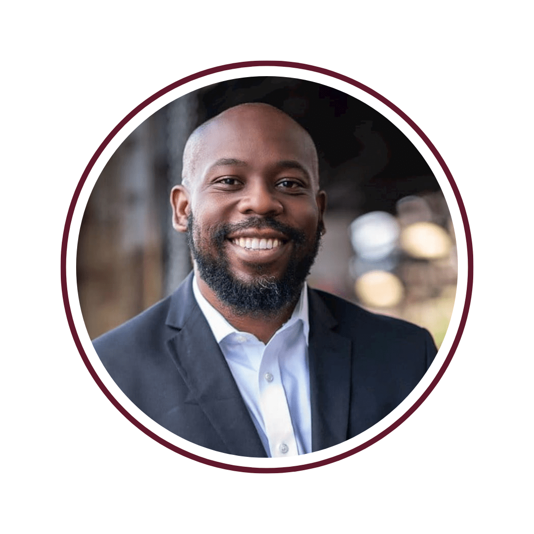 Young smiling man standing in front of a blurred background. You can see the glint of lights behind him. He is wearing a dark gray blazer and white undershirt. The picture has a maroon circle frame.  