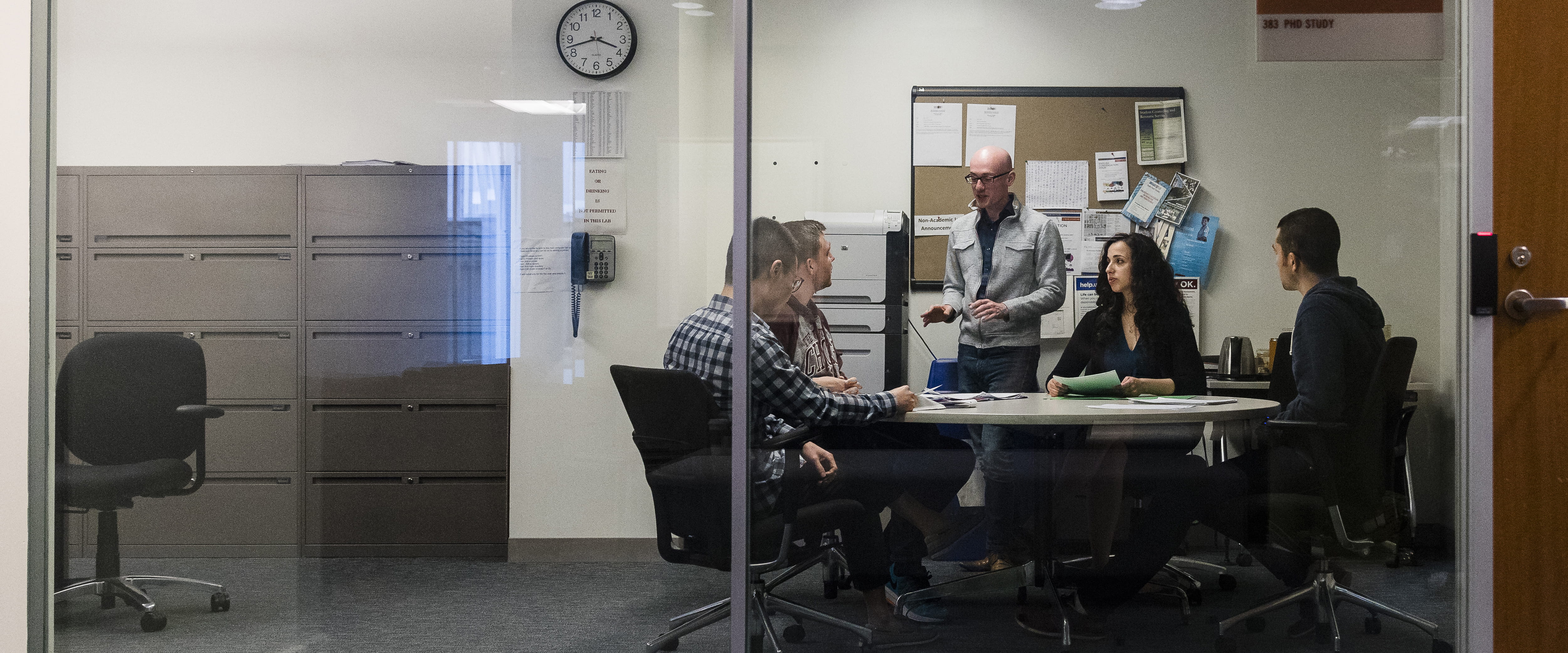 Group of four PhD students meet with their faculty advisor in study room with wall of windows.