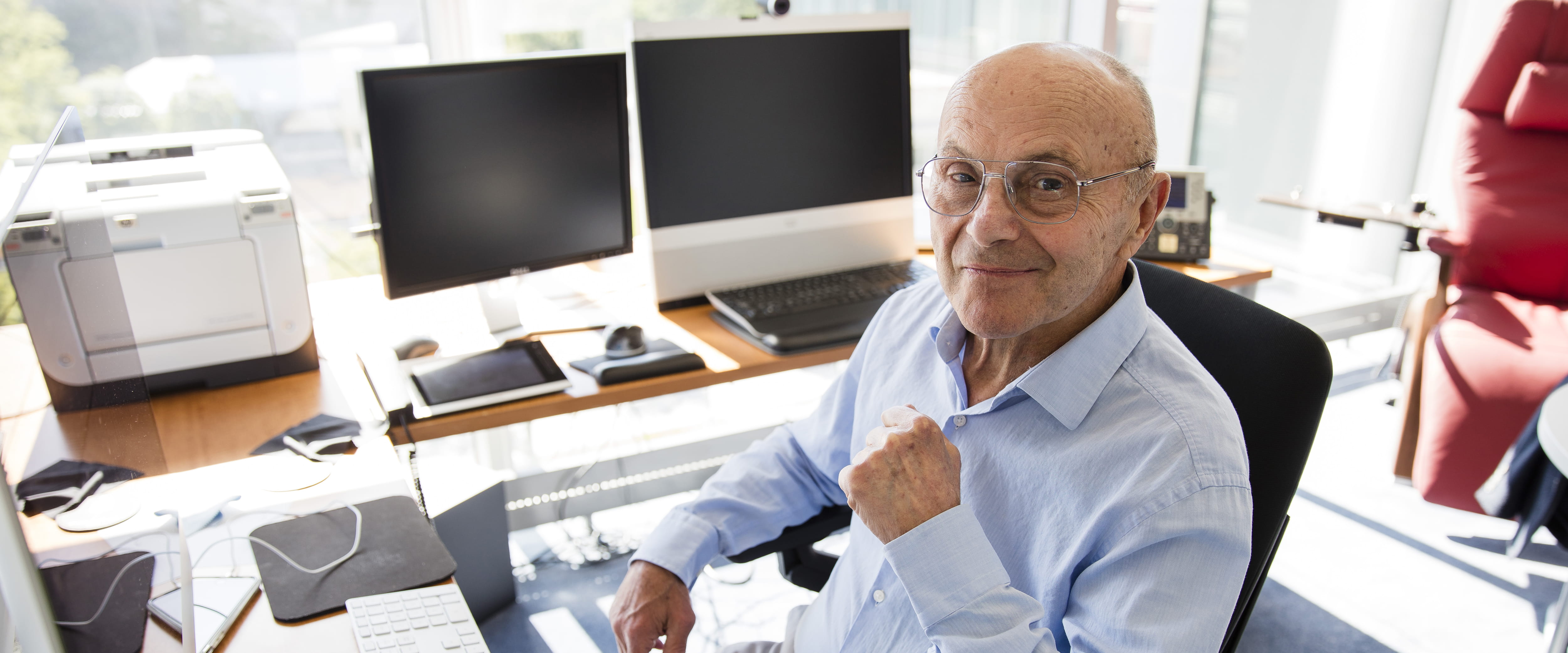 2013 Nobel Laureate in Economic Sciences Eugene Fama, an older man with button-down blue shirt, smiles while sitting at his desk.