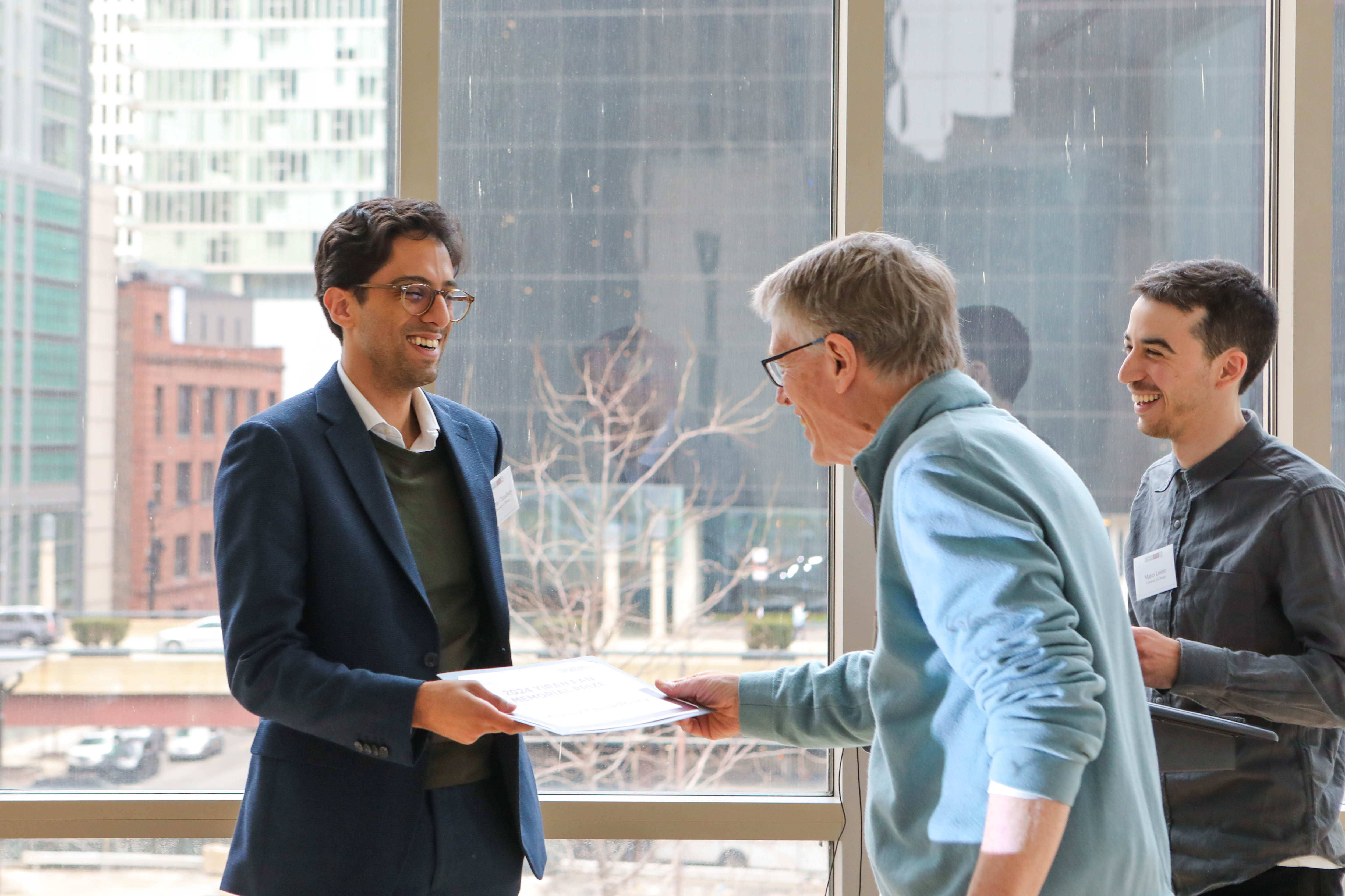 PhD student being handed an award by a faculty member in front of a glass window. Another student is smiling next to the awardee and award presenter. 