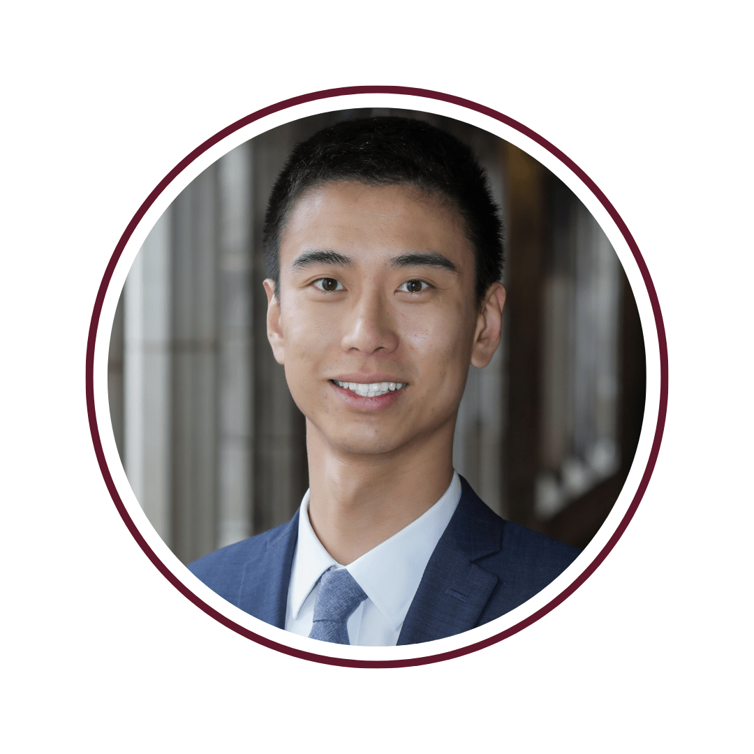 Young dark-haired man smiling in front of open corridor. He is wearing a blue suit jacket, blue tie, and a white undershirt. The picture is bordered by a maroon circle frame. 