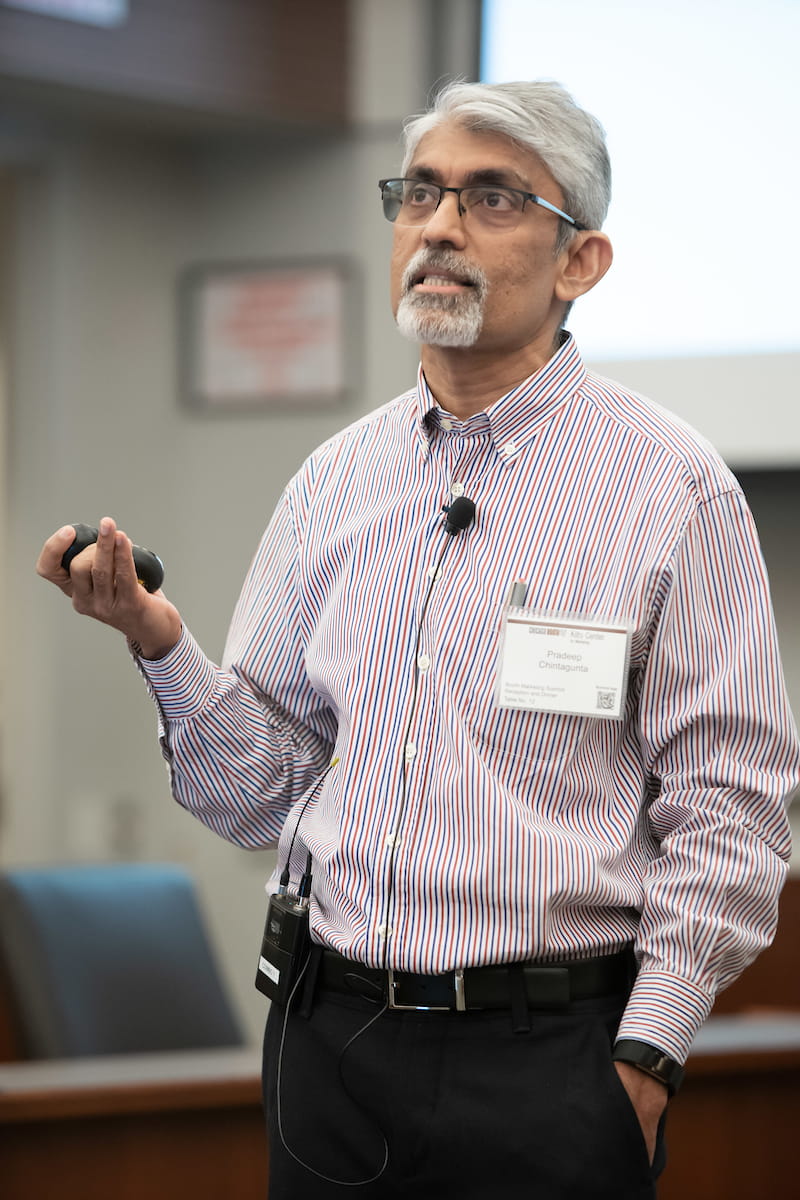 male professor standing and speaking, wearing glasses and a striped shirt