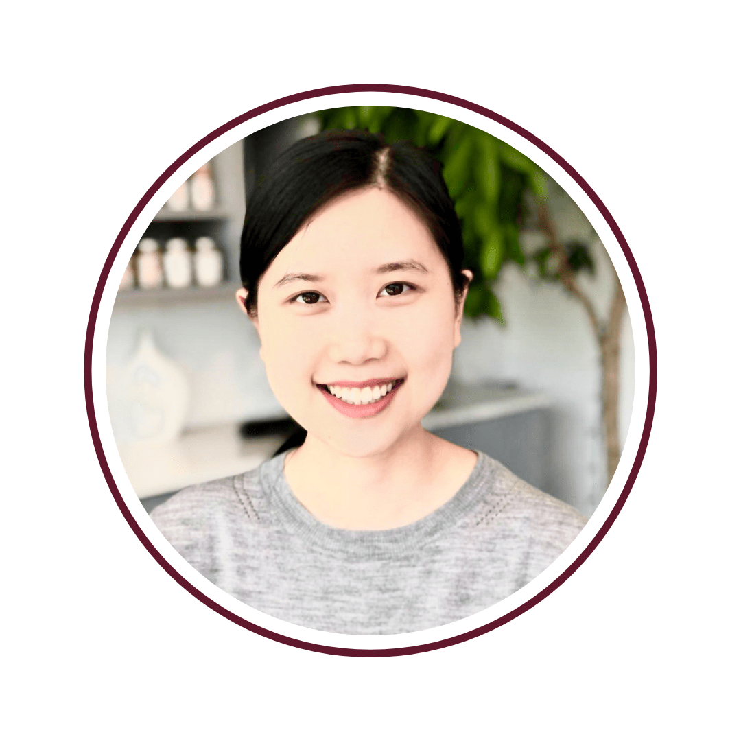 Picture of dark-haired, smiling woman in a grey shirt. The background is a tall, green plant and grey countertop. The picture is bordered by a maroon circle. 