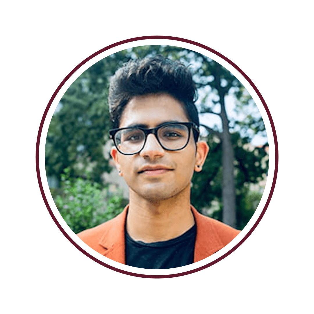 Picture of dark-haired young man wearing glasses, a black undershirt, and an orange blazer. He stands in front of tall, green trees. The picture is bordered by a maroon circle. 