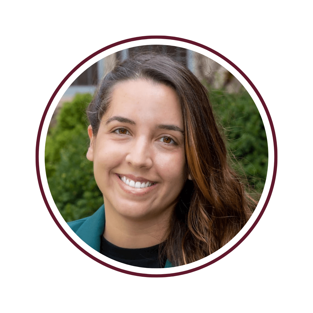 Young woman smiling in front of green foliage. She is wearing a teal blazer and black undershirt. The frame is bordered by a maroon circle. 