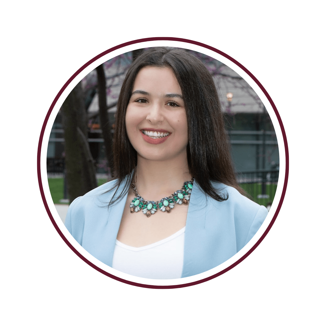 Smiling dark-haired woman standing in front of blooming tree with purple flowers. She is wearing a light blue blazer and a turquoise necklace. 