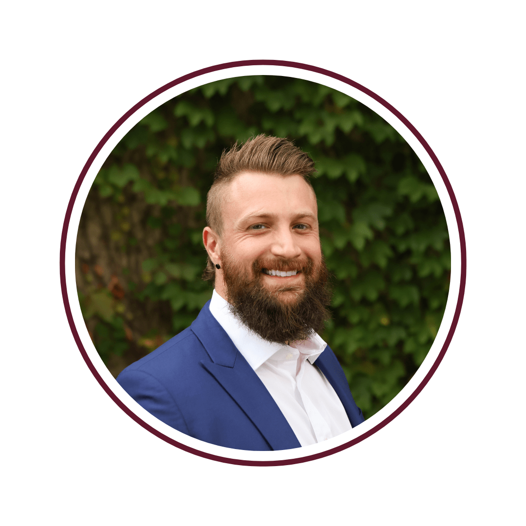 Headshot picture of blonde young man wearing a white dress shirt, navy blue blazer, and red tie. The background is grey. The picture is bordered by a maroon circle. 