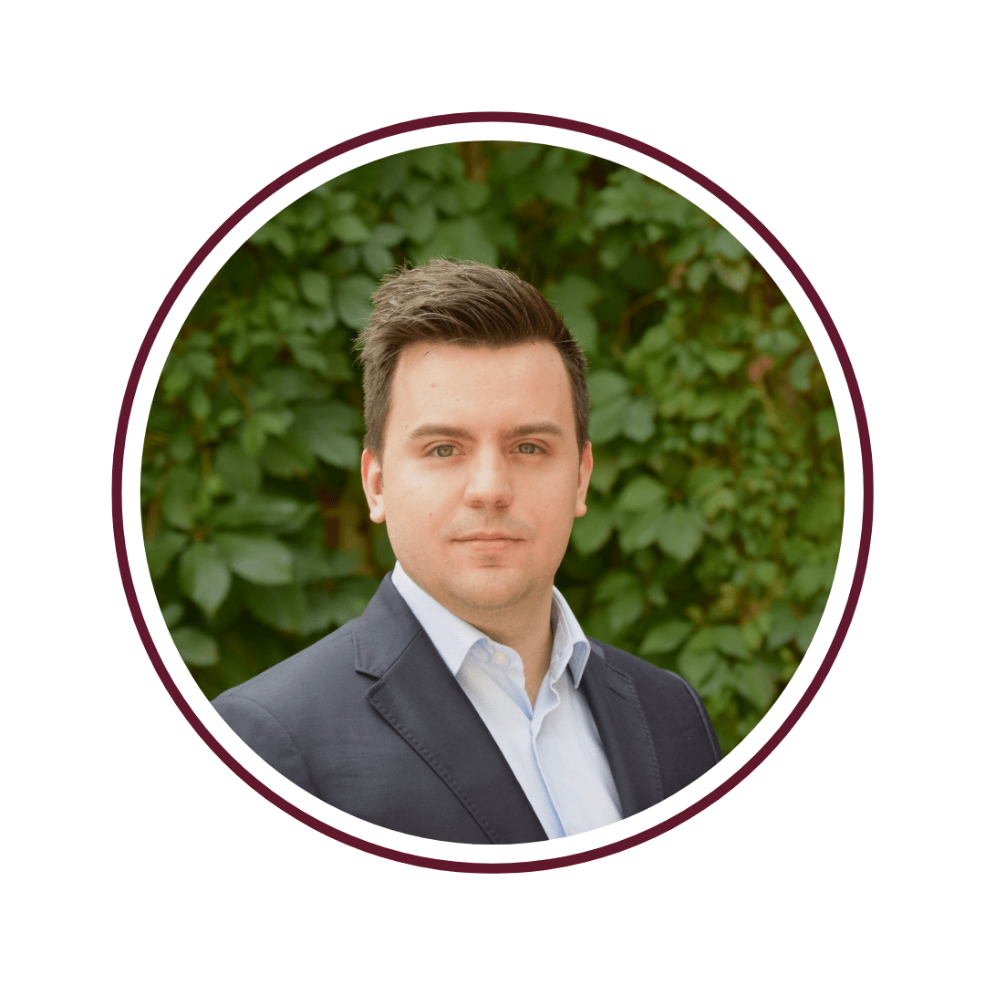 Picture of dark-haired man wearing a black blazer and blue dress shirt who is standing in front of a green vine background. The picture is bordered by a maroon circle. 