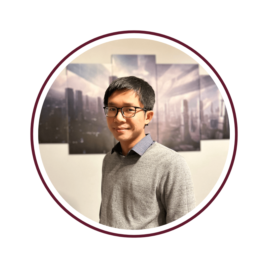 Headshot of young, smiling, dark-haired man wearing glasses and a grey long-sleeved shirt. He stands in front of a five-panel photograph of a city skyline. The photo is bordered by a maroon circle. 
