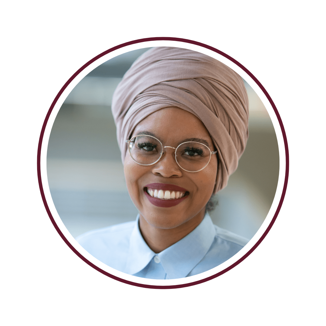 Picture of smiling young woman wearing a light pink turban, glasses, and blue dress shirt. 