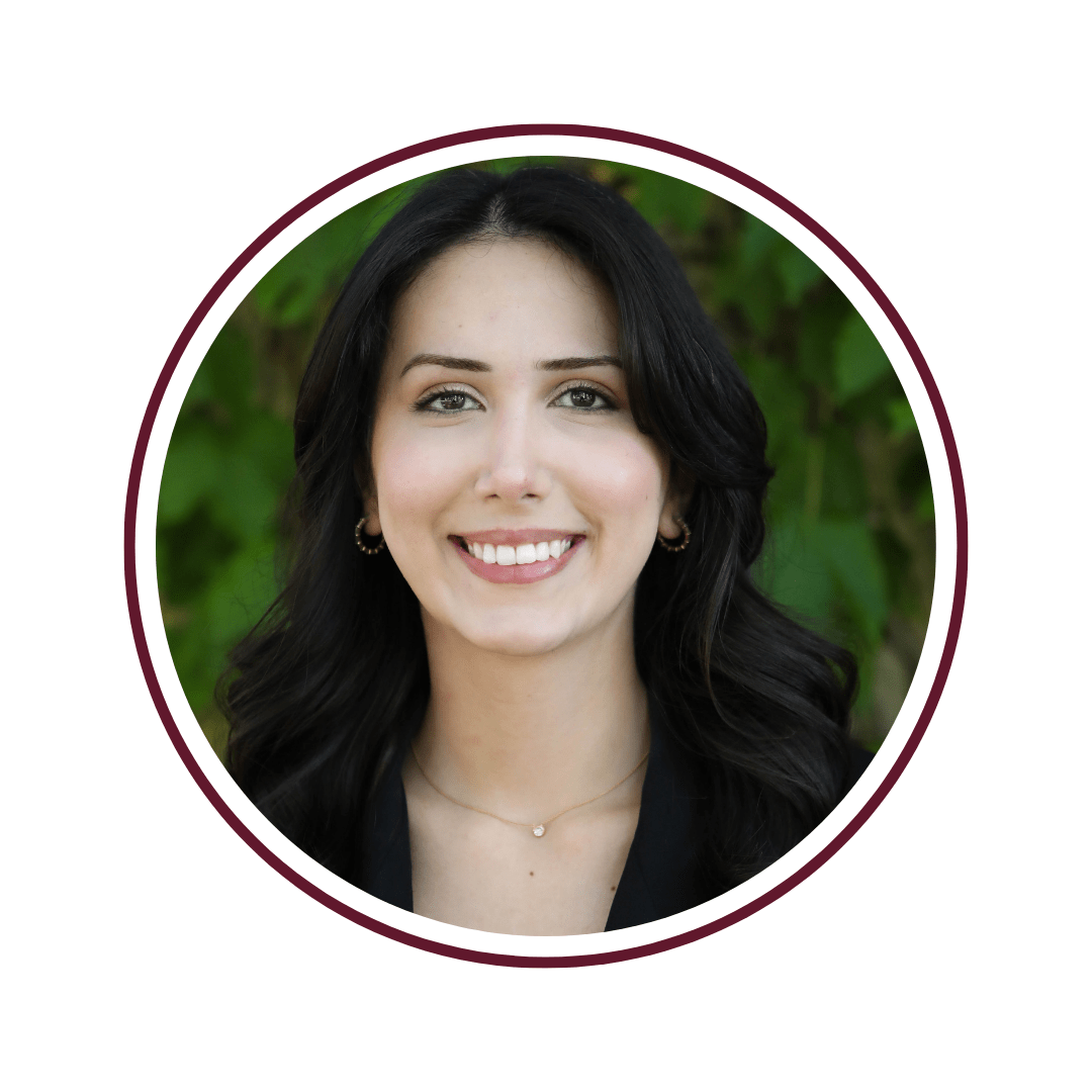 Picture of young dark-haired woman wearing a white blouse. She is smiling in front of a stretch of tall, green trees. The blue sky is visible overhead. 