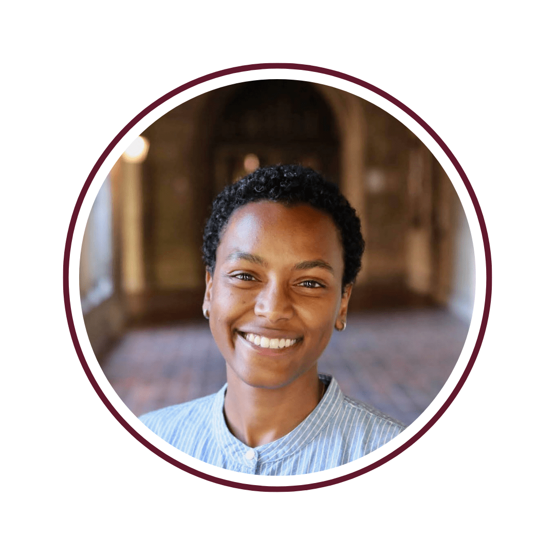 Picture of smiling woman with curly hair and wearing glasses. She is standing in front of a green-vined background. 