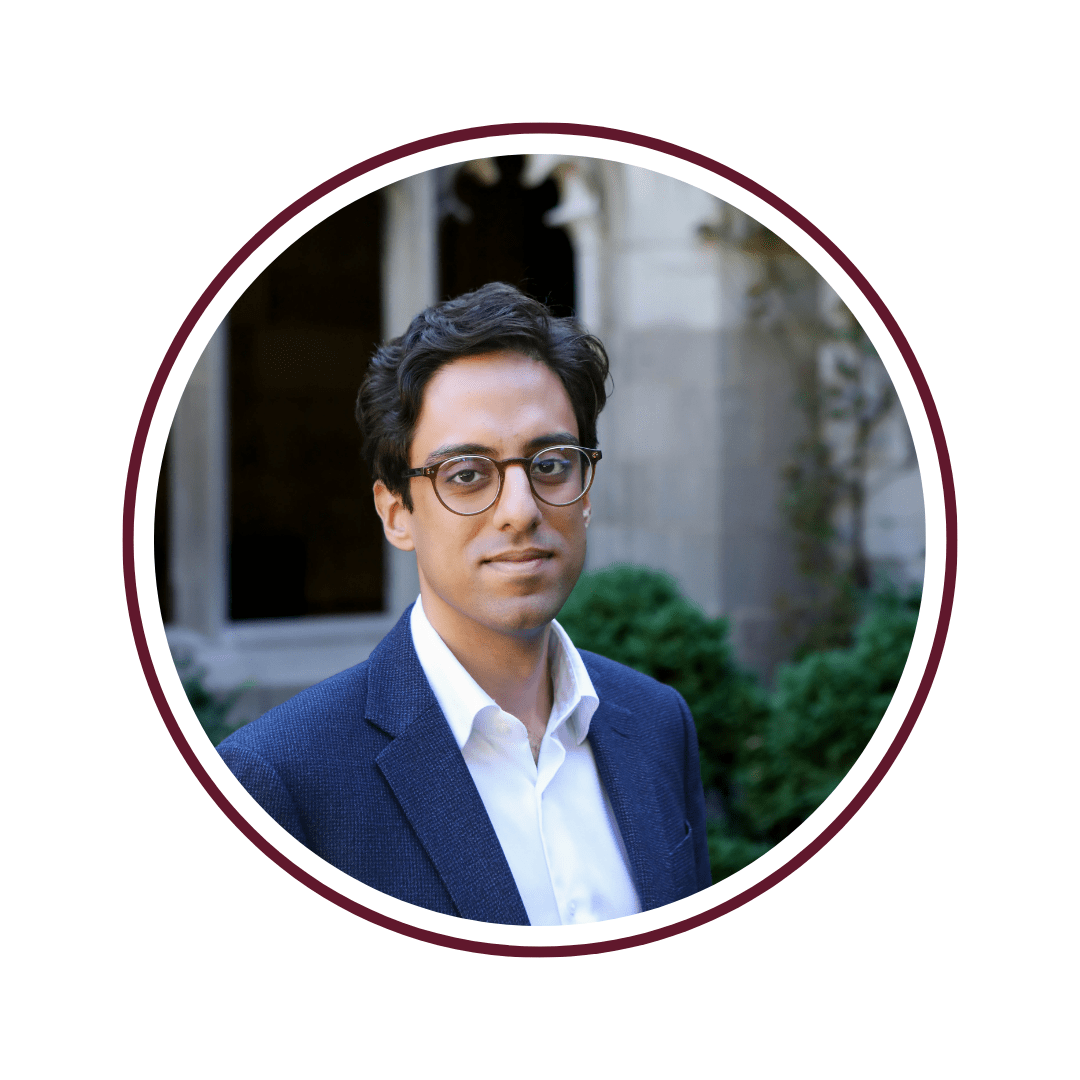 Picture of young dark-haired man wearing glasses and a grey blazer. He is standing in front of a off-white background with crown moulding. 