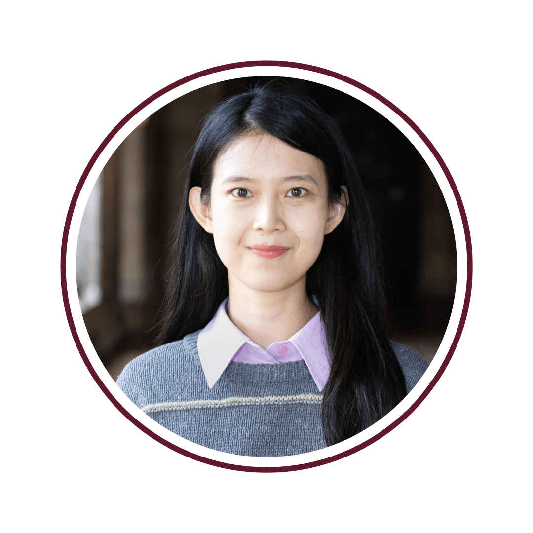 Picture of dark-haired woman in blue and green sweater with a light purple dress shirt underneath. She is standing in front of a row of column windows. The picture is bordered by a maroon circle. 