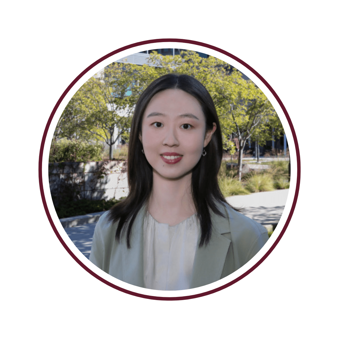 Headshot of smiling, dark-haired woman wearing a cream-colored blouse and a grey blazer. The background is of the external view of the Chicago Booth Harper Center, with trees and the building's rear facade visible. The picture is bordered by a maroon circle. 