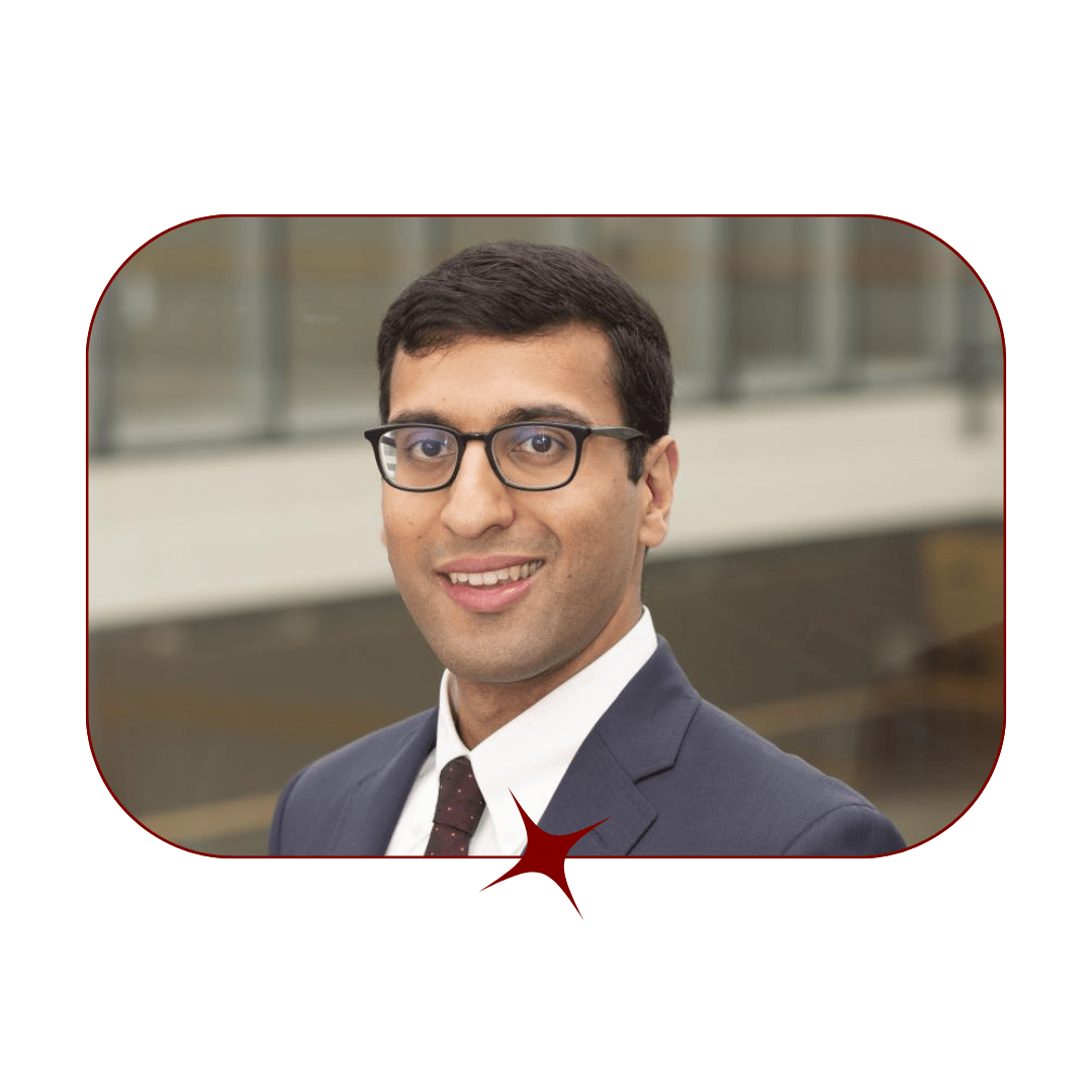 Picture of a young, smiling, dark-haired man wearing glasses and a grey suit and tie in front of a blurred wooden background. The picture is bordered by a thin, red-lined frame with a red star in the center. 