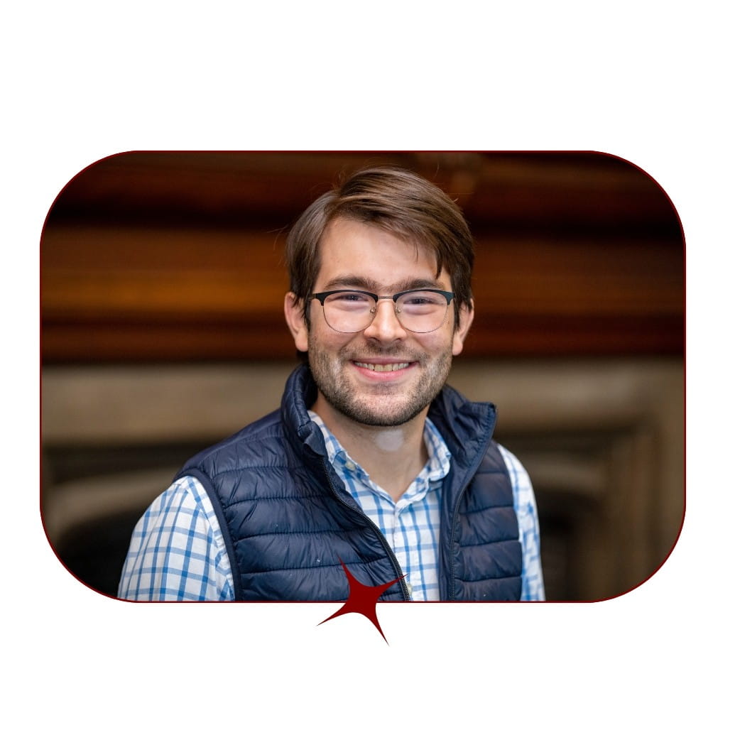 Picture of a young, smiling, dark-haired man wearing glasses and a blue, checkered shirt and vest in front of a blurred wooden background. The picture is bordered by a thin, red-lined frame with a red star in the center. 