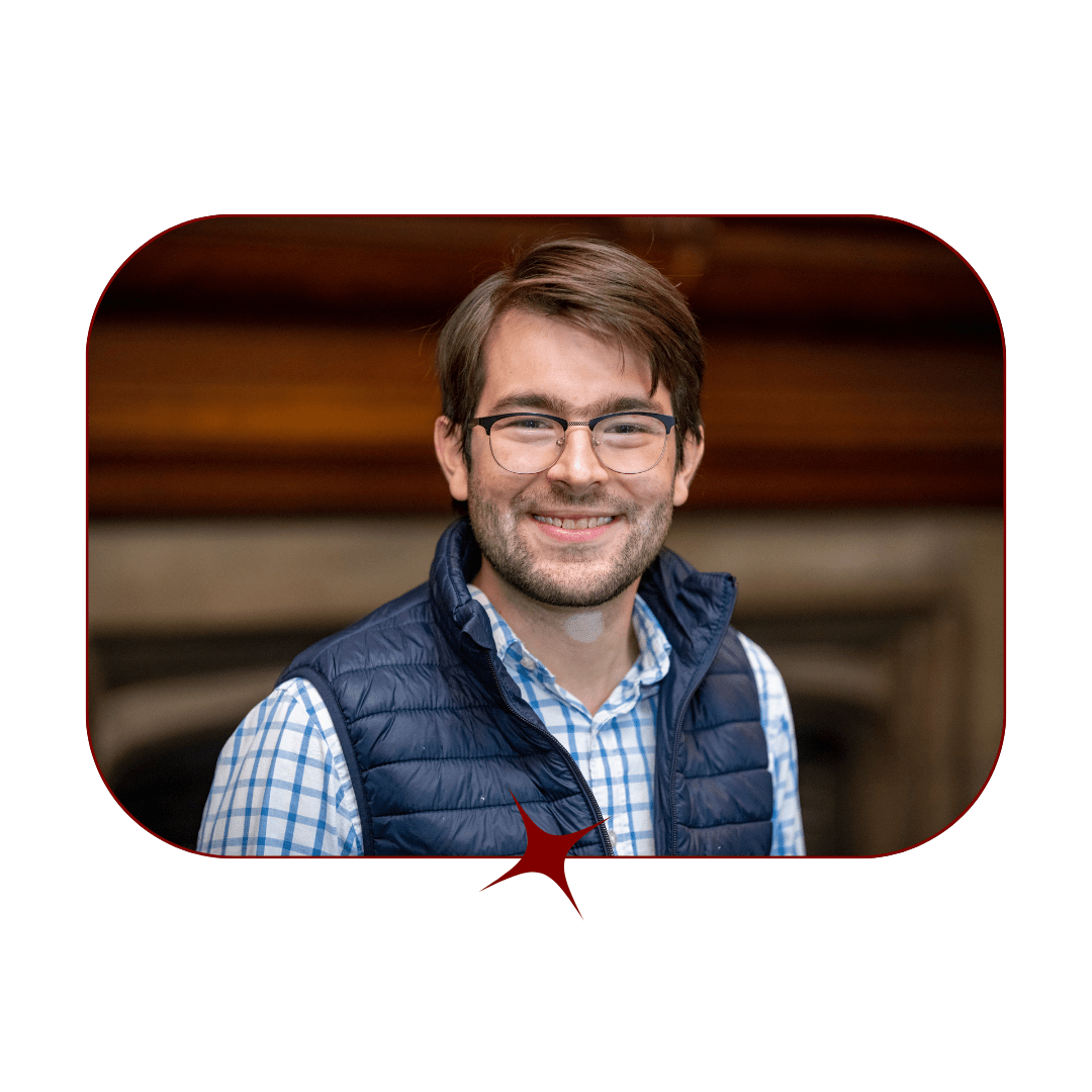 Picture of a young, smiling, dark-haired man wearing glasses and a blue, checkered shirt and vest in front of a blurred wooden background. The picture is bordered by a thin, red-lined frame with a red star in the center. 