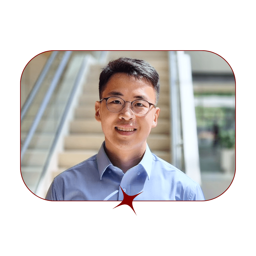 Picture of a young, smiling, dark-haired man wearing glasses and a blue button-up in front of a marble staircase. The picture is bordered by a thin, red-lined frame with a red star in the center. 