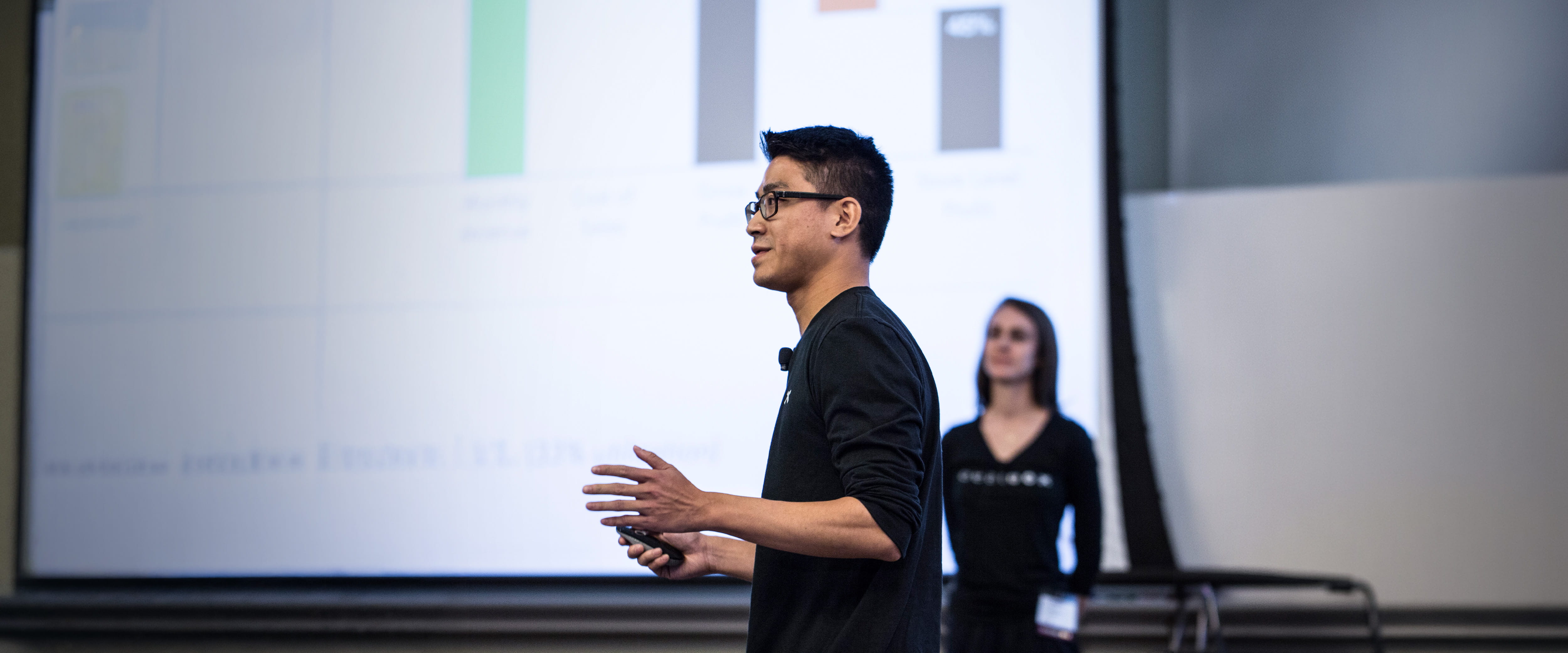 A student stands in front of a large presentation while gesturing with his hands.