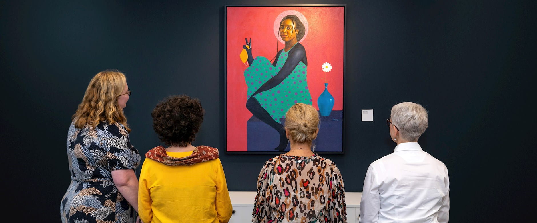 Four women look at a painting of a woman with a halo holding an orange face mask at the London Conference Centre