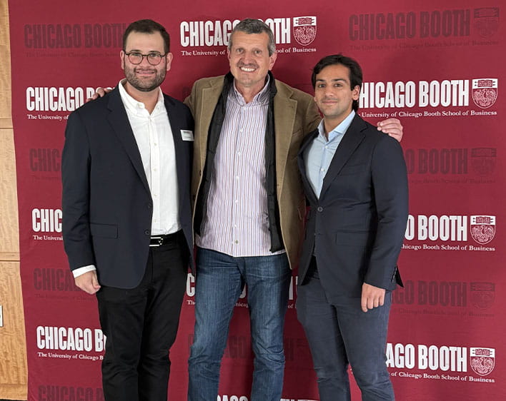 Christos Papandreou with two individuals smiling in front of Chicago Booth banner