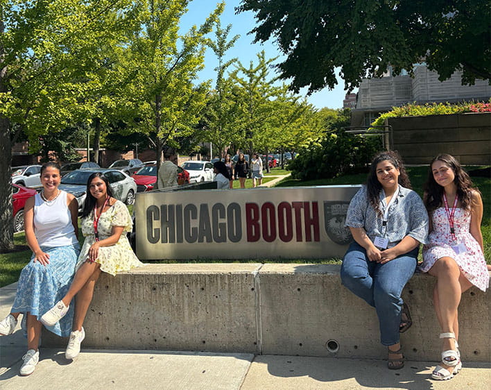 Nehir Mentes and three other Boothies smiling while sitting down near Chicago Booth sign outside of the Harper Center