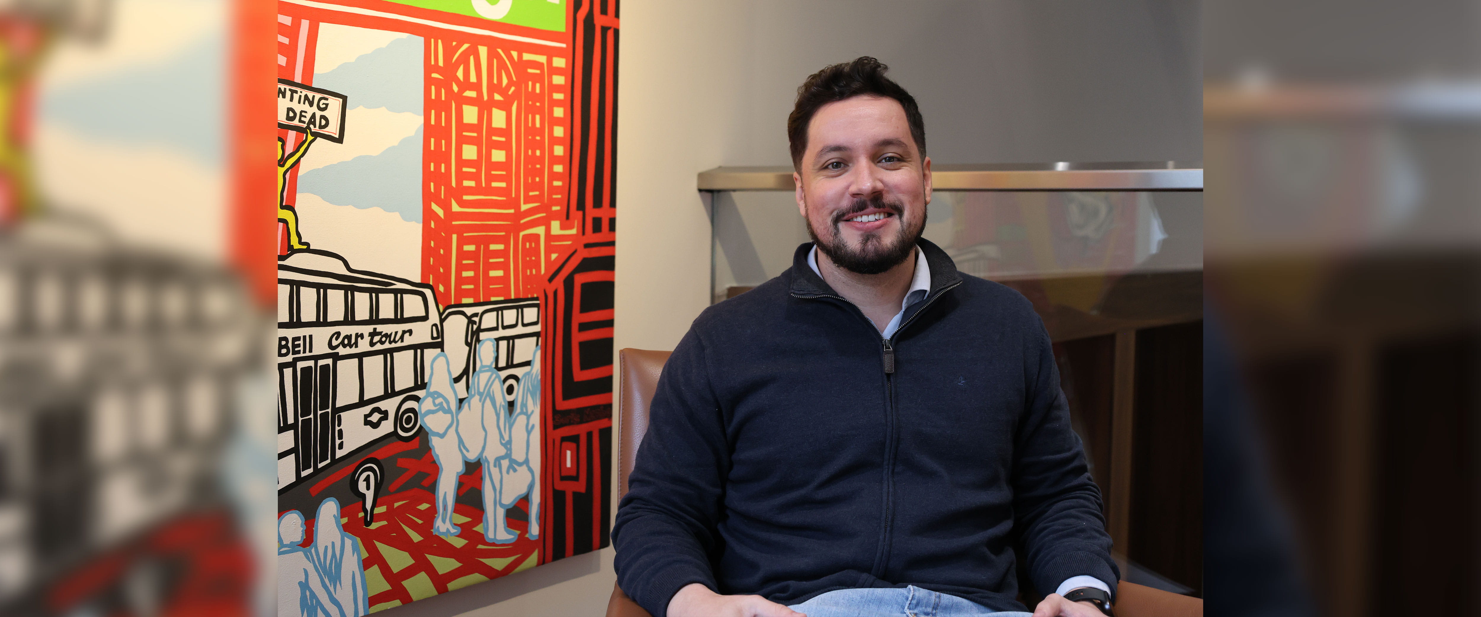 Caio smiling while sitting down in chair in front of artpiece 
