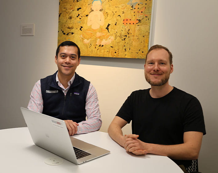 Booth students, Philipp Gschoepf and John Fabros sitting down at table with laptop smiling